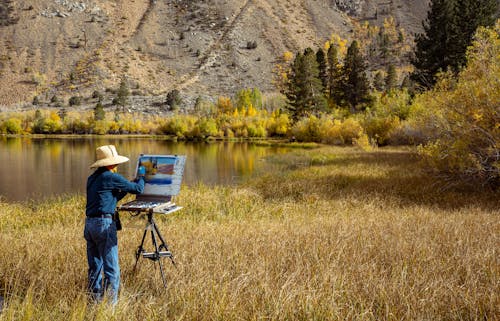 A Man in Sun Hat Standing on Brown Field while Painting on a Canvas
