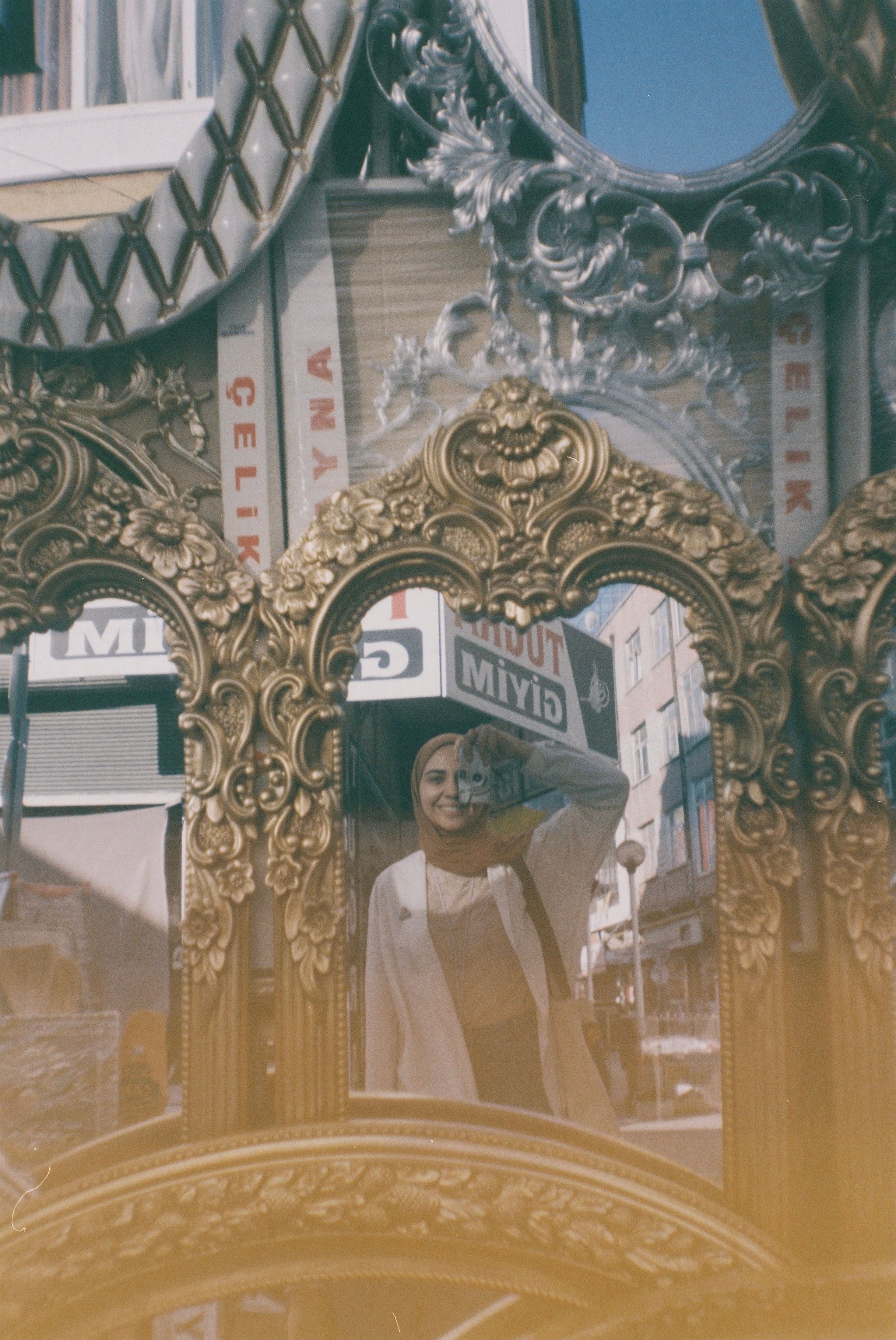 A woman captures a selfie in an intricate mirror with ornate golden frames, reflecting whimsical urban surroundings.