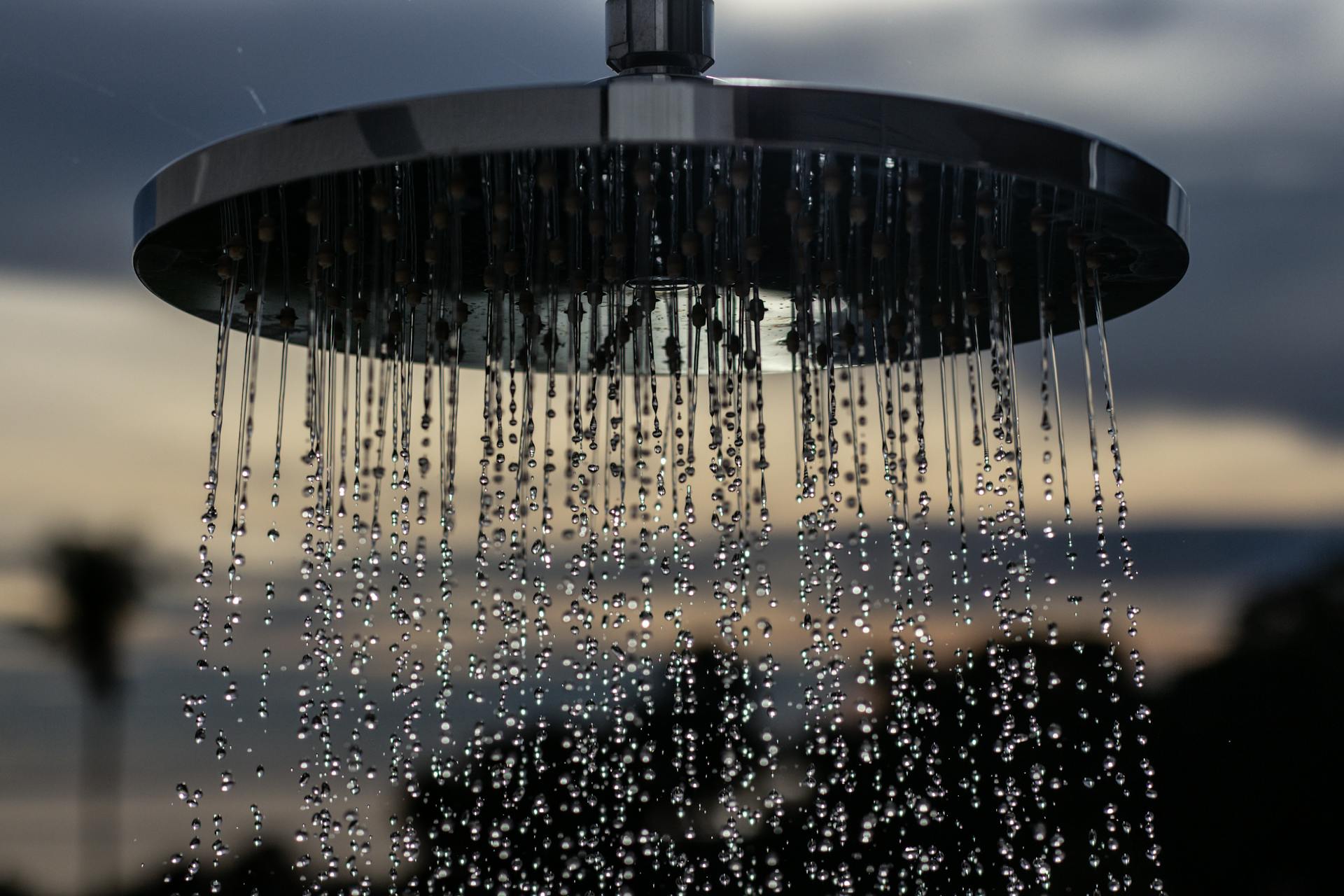 A detailed view of a shower head outdoors with drops flowing and a blurred sunset background.