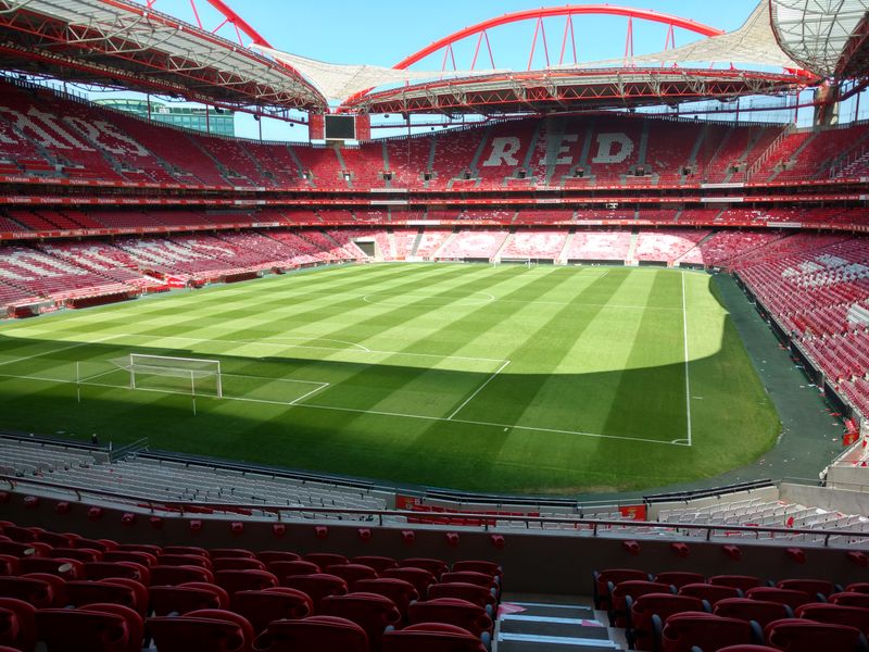 Inside the empty Estádio da Luz in Lisbon: red seats around a sunlit pitch under the red roof arches.