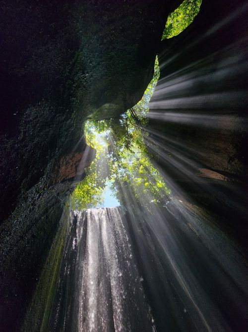 Free Sunlight beams through a cave crevice, illuminating a cascading waterfall in a stunning natural scene. Stock Photo