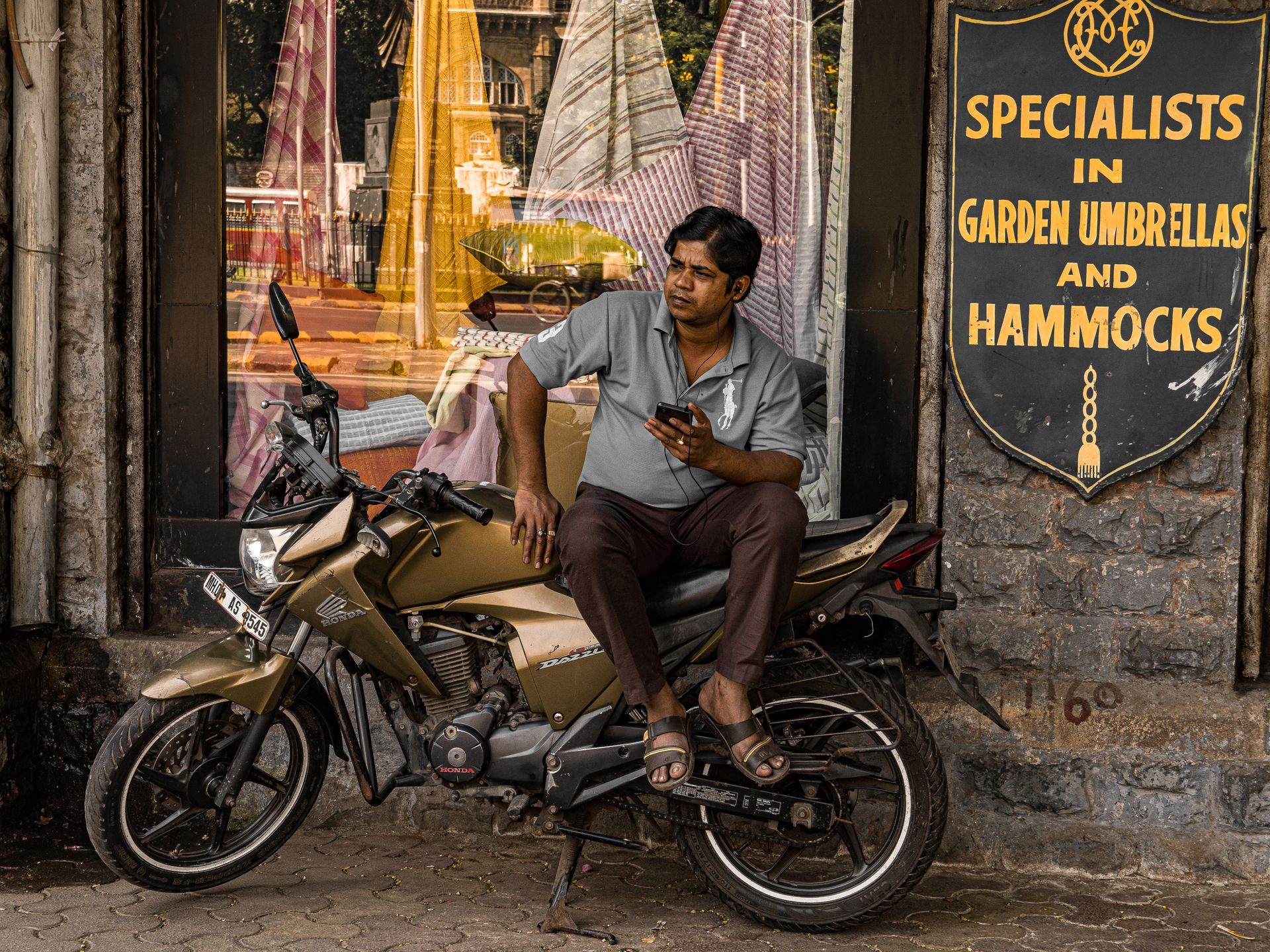A South Asian man sits on a parked motorcycle in Mumbai, India, using his phone in front of a storefront.