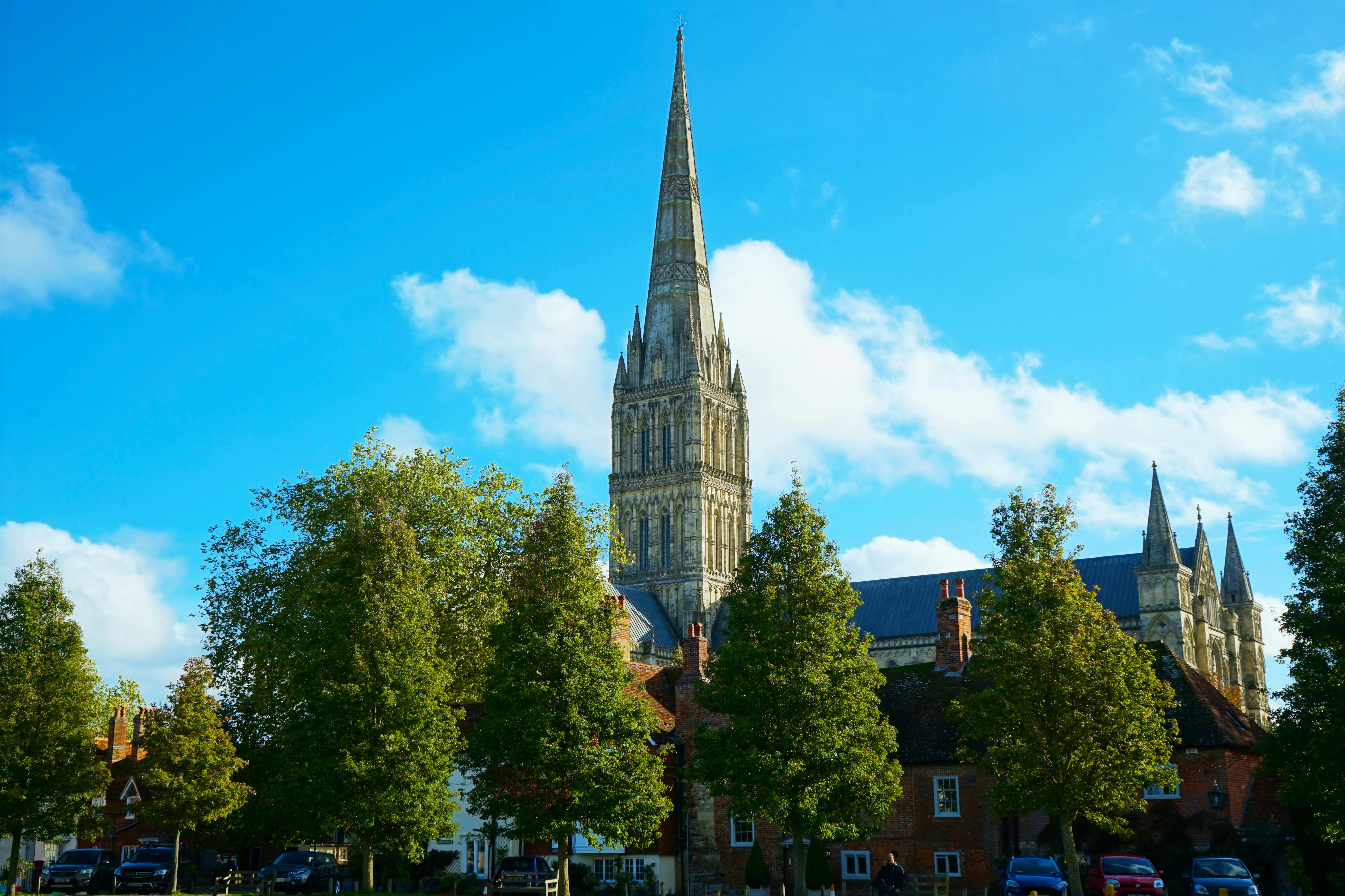Elevated view of the iconic Salisbury Cathedral spire surrounded by lush trees under a bright blue sky.