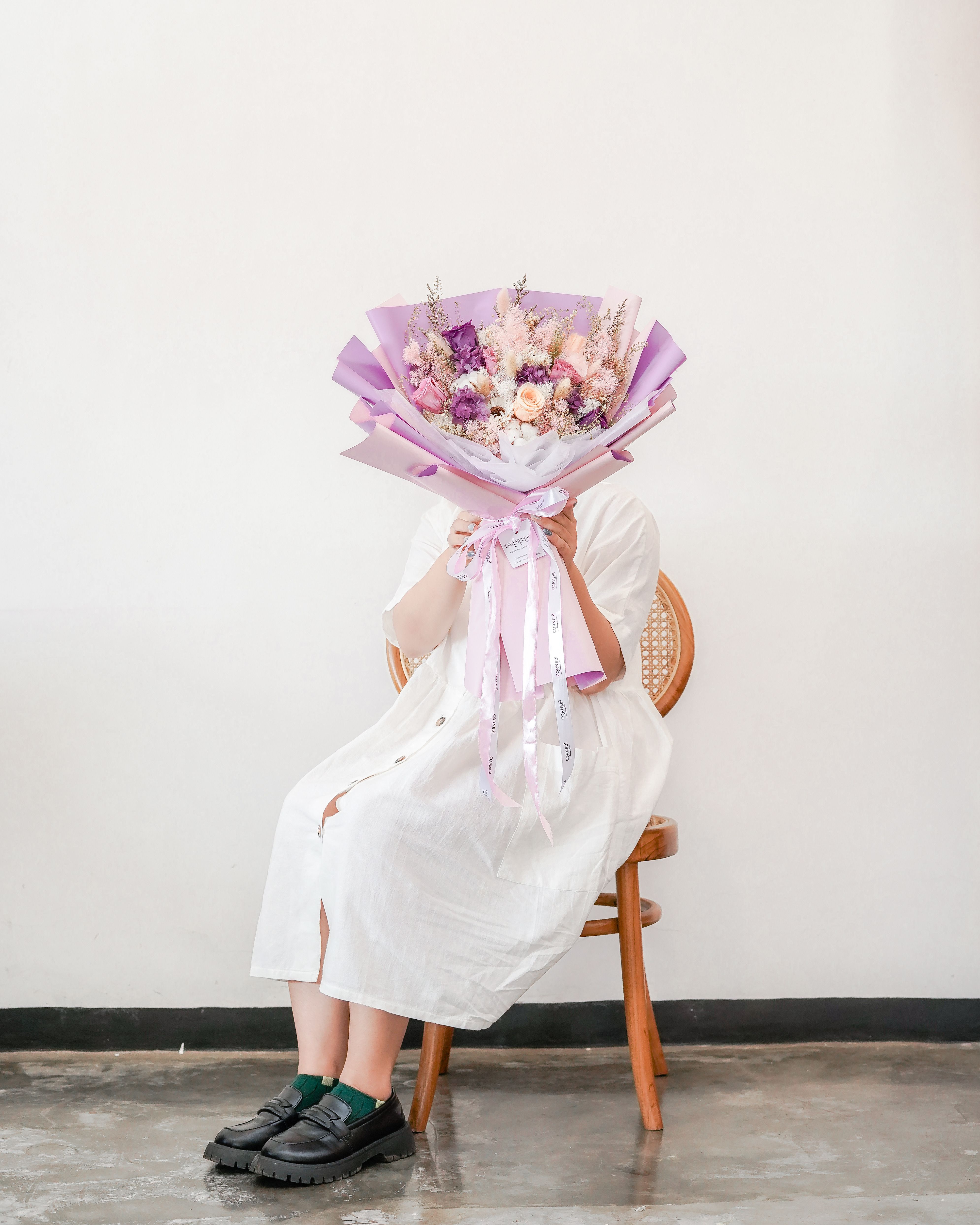 A person in a white dress sits on a chair holding a colorful flower bouquet indoors.