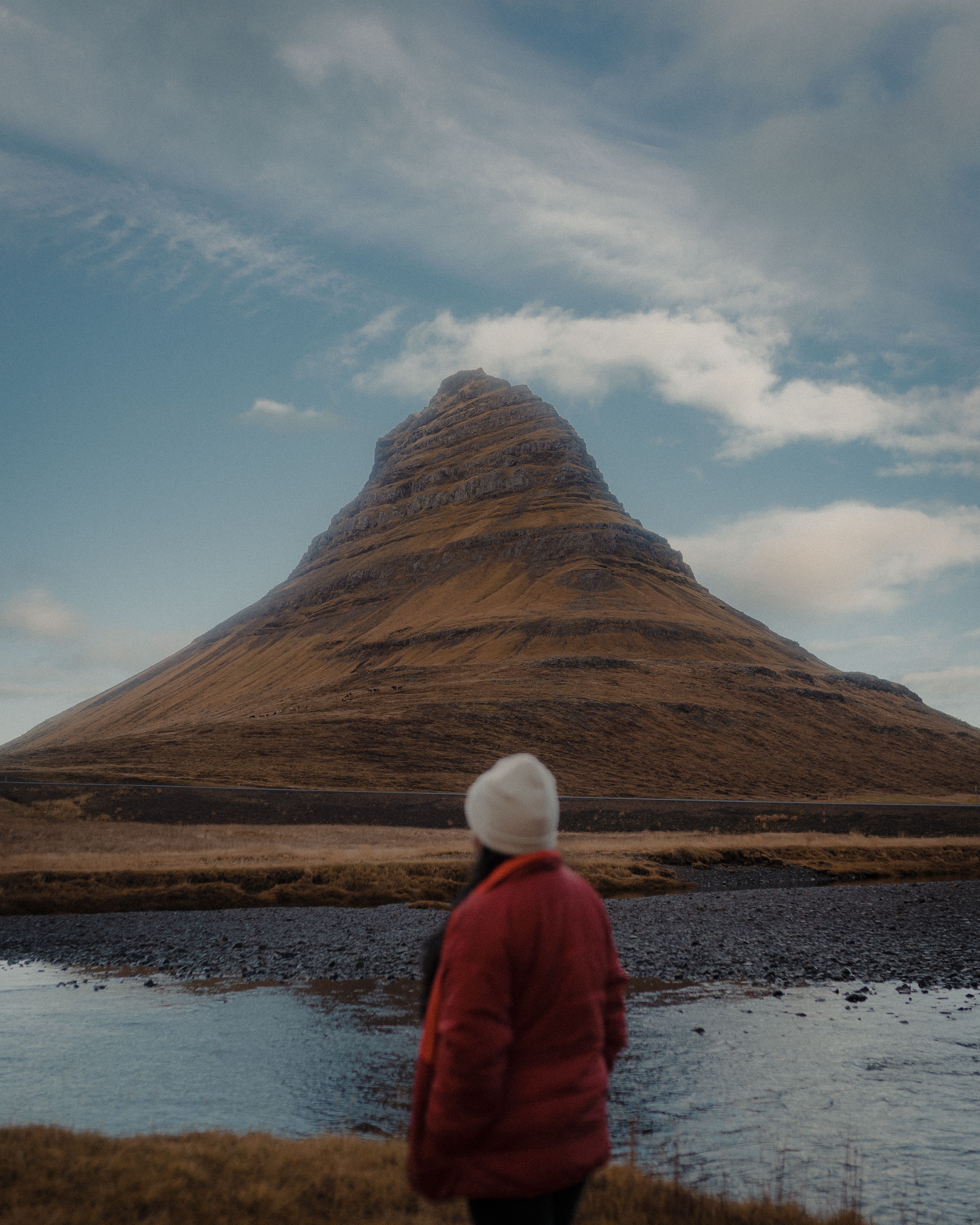 A woman in a red jacket stands facing a cone-shaped mountain by the lakeshore, capturing the raw beauty of nature.