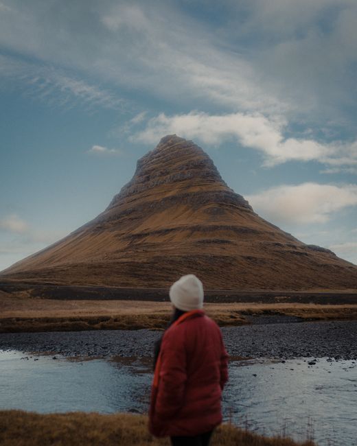 A woman in a red jacket stands facing a cone-shaped mountain by the lakeshore, capturing the raw beauty of nature.