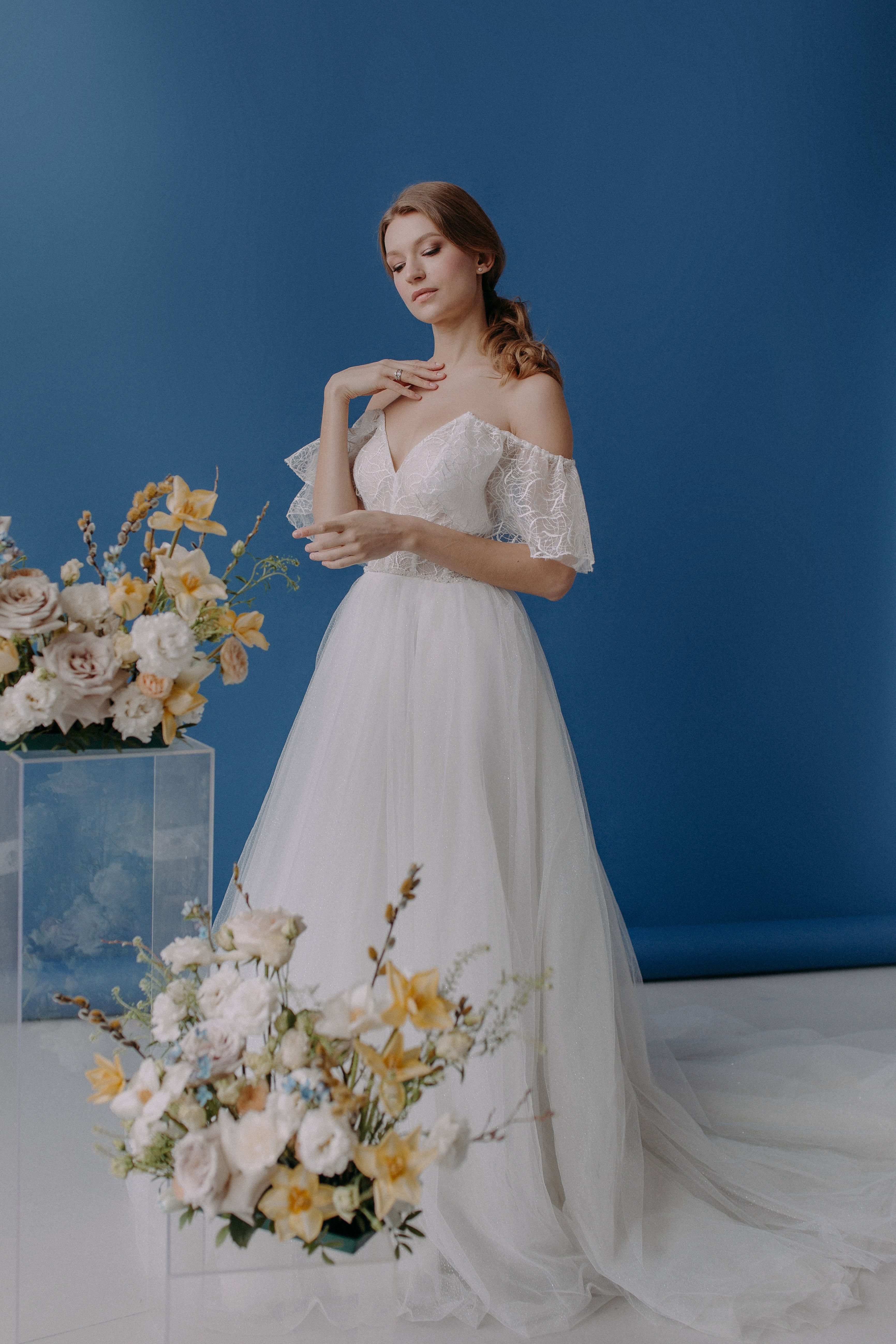 A bride in a white gown poses gracefully with floral arrangements against a blue backdrop.