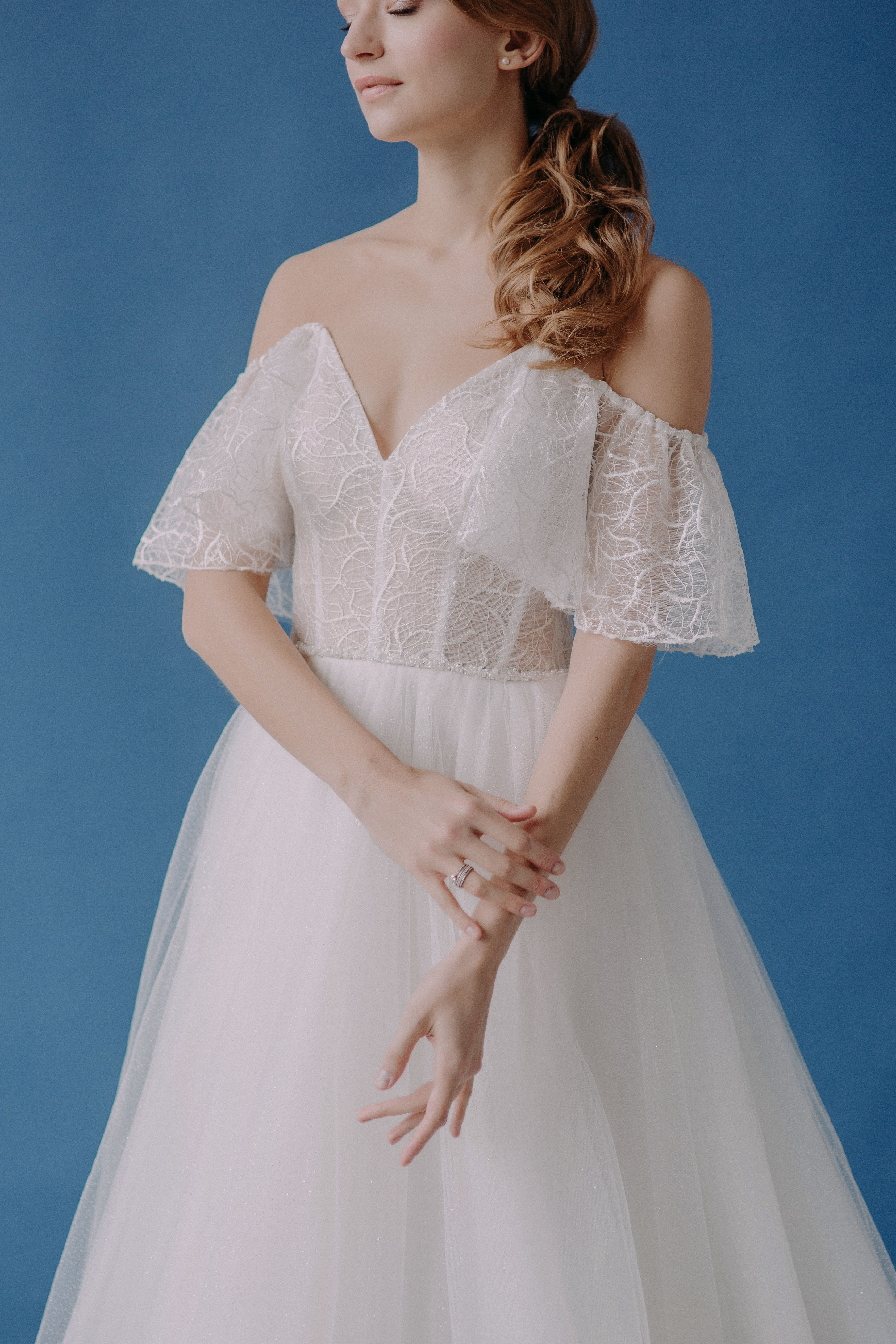 Portrait of a woman in a lace off-shoulder white gown against a blue background.
