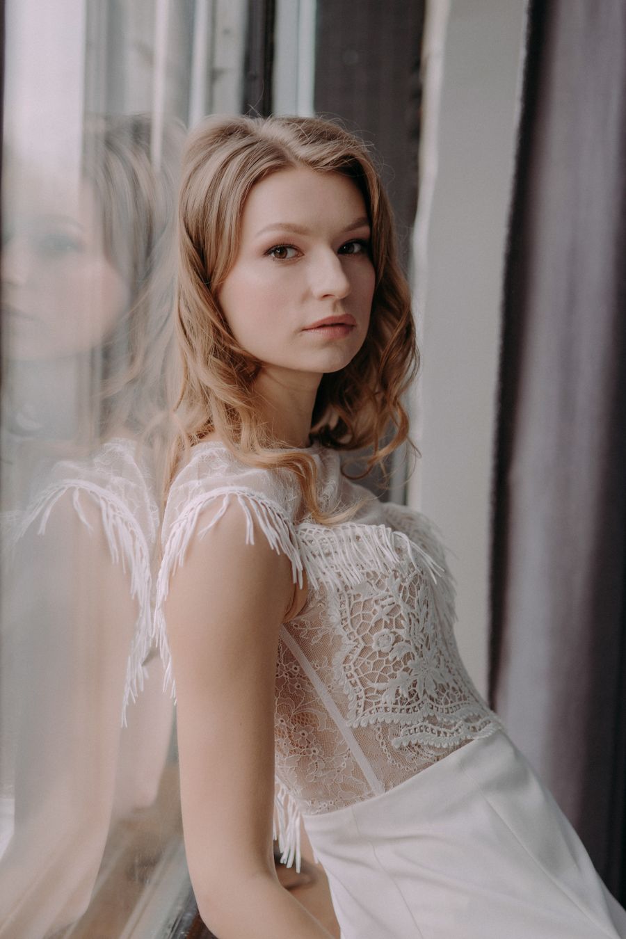 Woman in an intricate white lace dress standing by a window