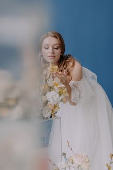 A graceful woman in a white gown enjoying the fragrance of flowers against a blue backdrop.