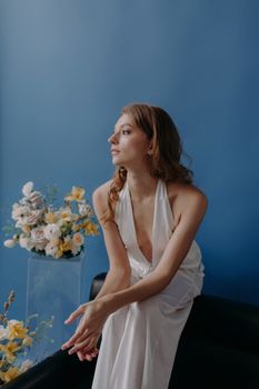 Elegant woman in a white dress posing gracefully with floral arrangement in a studio.