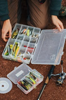 Fisherman arranging fishing lures in a tackle box beside a fishing rod.