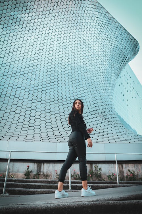 Woman in Black Jacket and Black Pants Standing Near White Concrete Building with Geometric Design