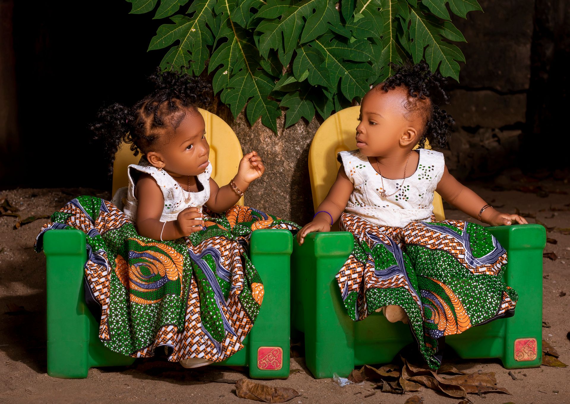 Charming twin toddlers sitting outside in vibrant traditional dresses, sharing a playful moment.