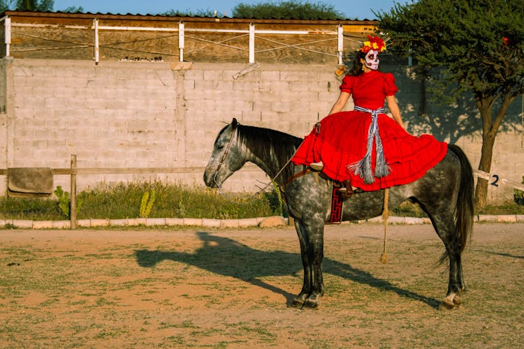 A Skull Face Painted Woman Wearing A Red Dress Riding A White And Black Horse