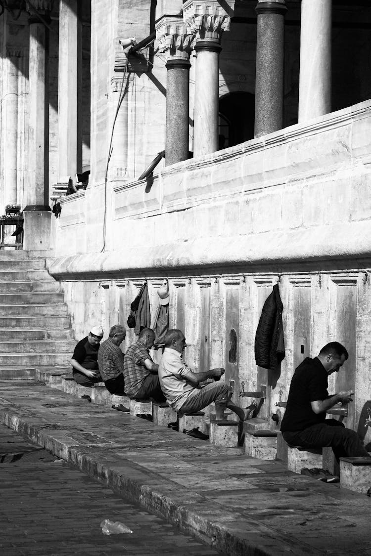 Grayscale Photo Of Men Sitting In Front Of Faucets Outside A Mosque