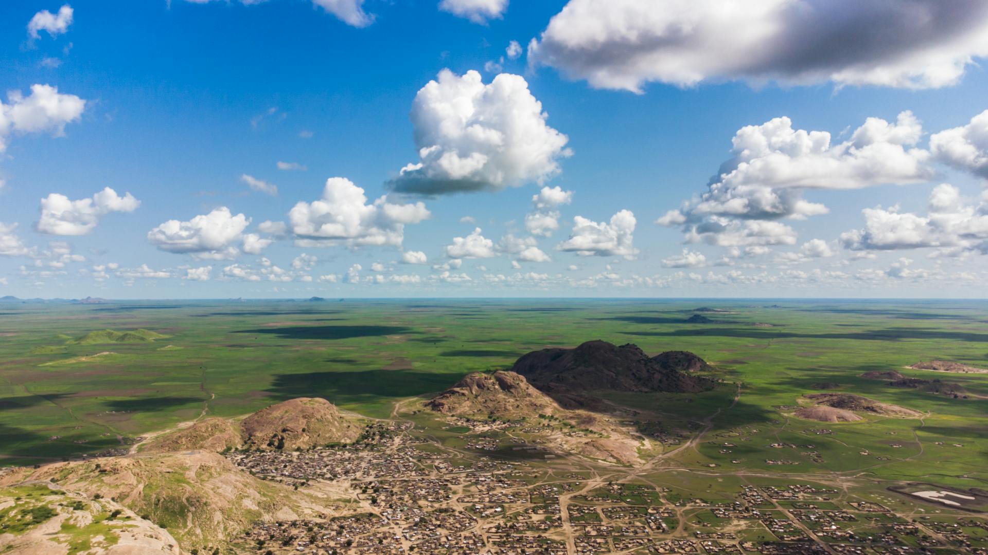 Stunning aerial shot of Baylah, Sudan showcasing hills, fields, and townscape under a blue sky with clouds.