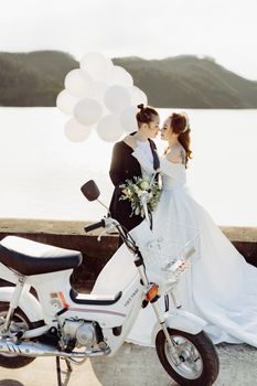 A lovely lesbian wedding couple sharing a tender moment by a scenic lake, featuring a white scooter and balloons.