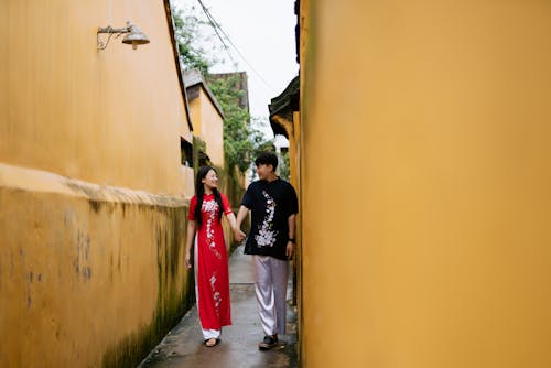Man in Black Shirt Walking Hand in Hand with a Woman in Red Ao Dai in an Alley 