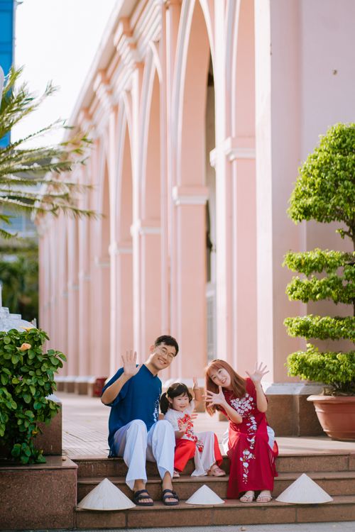 Free A cheerful family of three sitting outdoors near a pink building, smiling and waving. Stock Photo