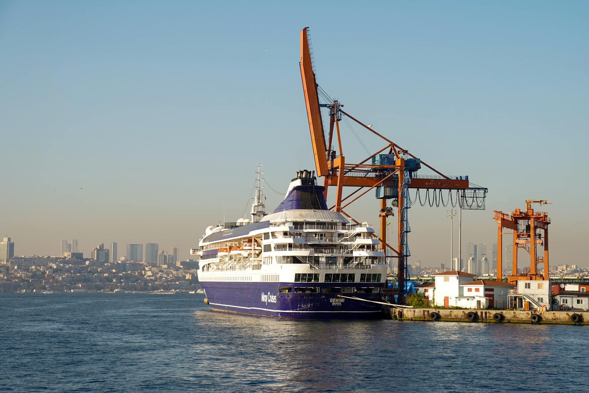 A majestic cruise ship docked at a busy port with cargo cranes under a clear blue sky.