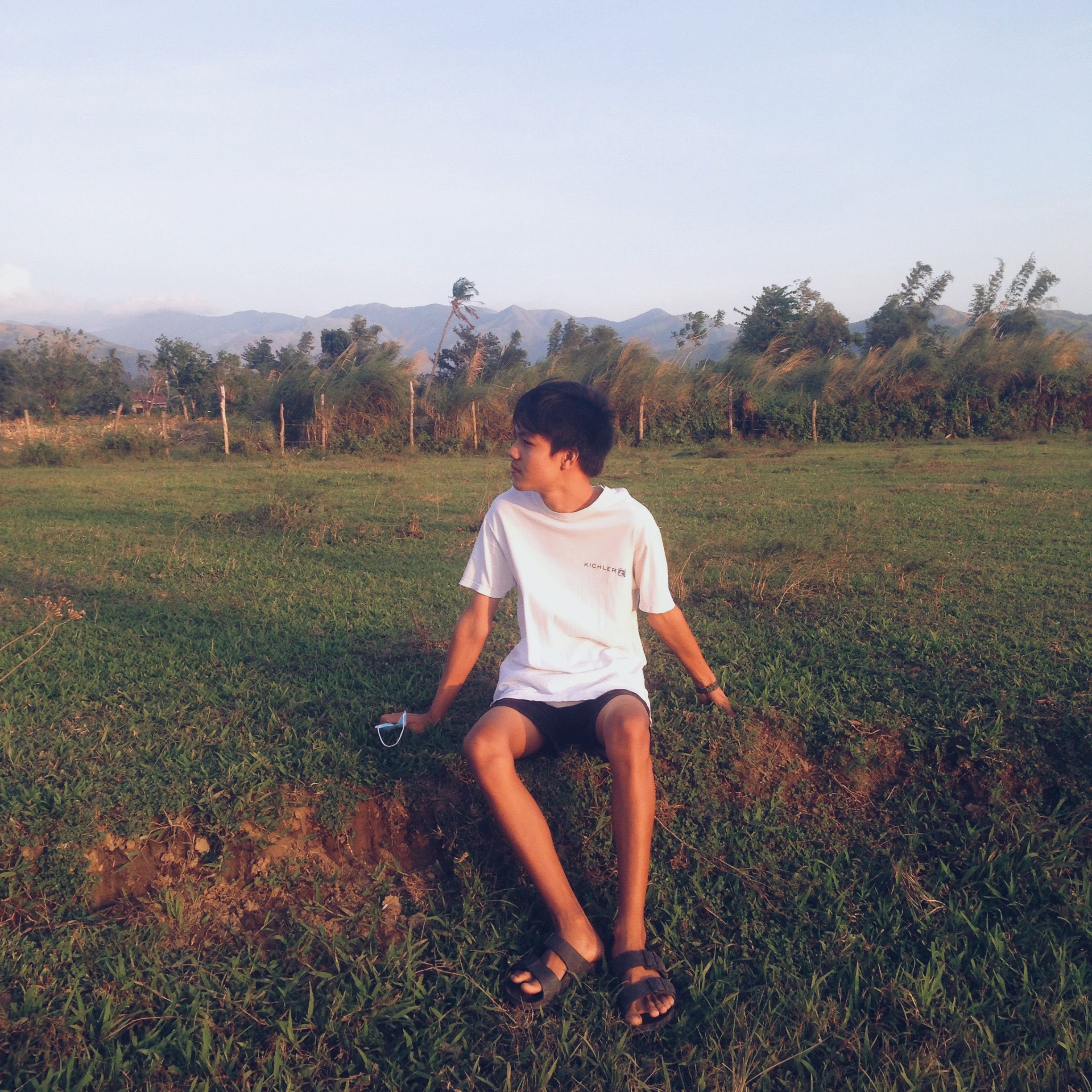 A teenage boy sits in a grassy field at sunset with mountains in the background.