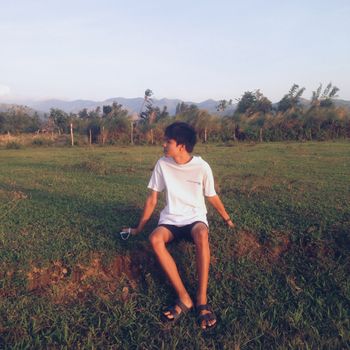 A teenage boy sits in a grassy field at sunset with mountains in the background.