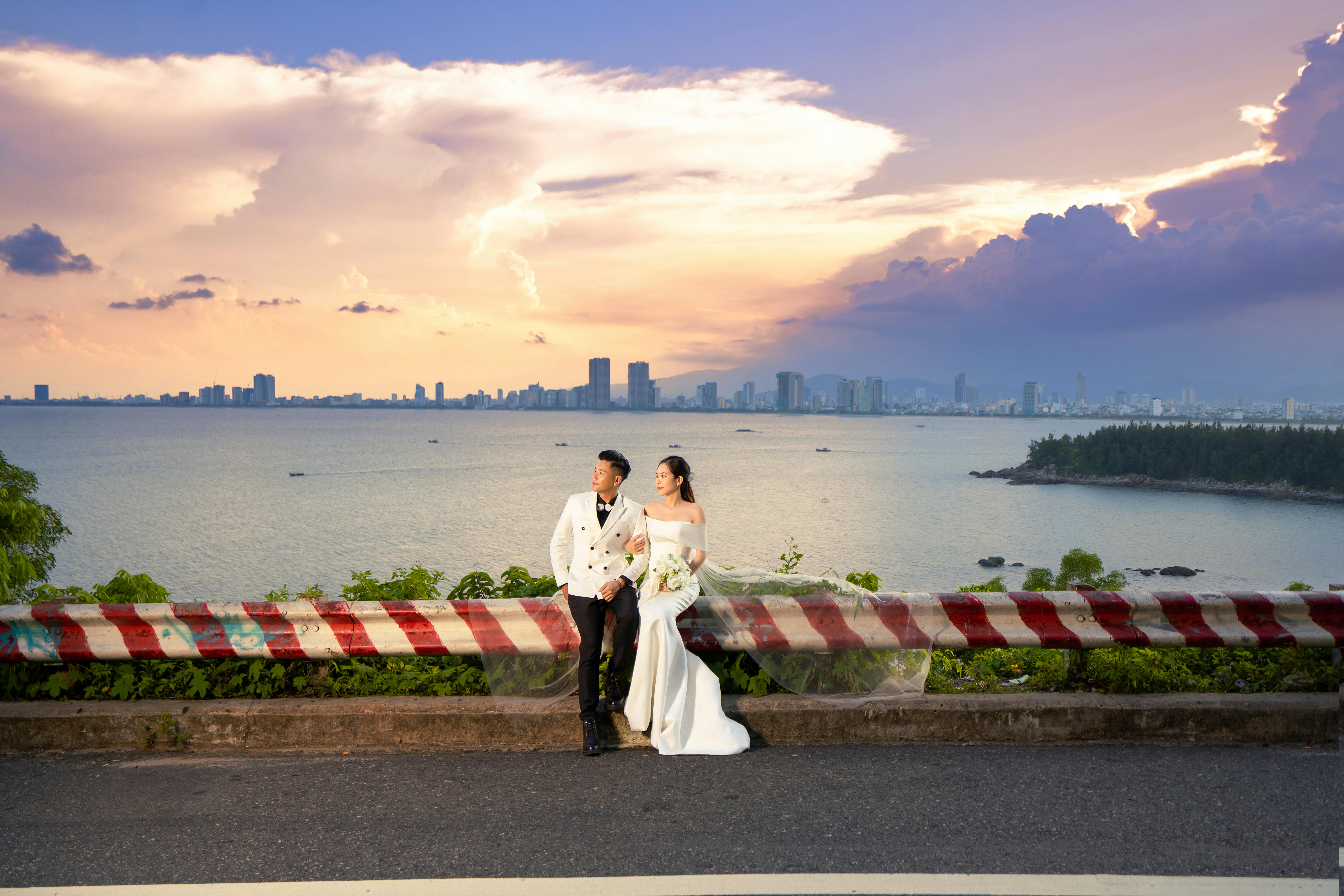 Newlywed couple in elegant attire enjoying a scenic sunset view by the ocean city skyline.