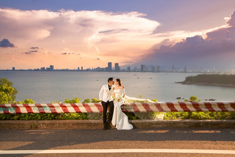 A Bride And Groom Sitting At The Road Barrier Near The Ocean While Doing Face To Face