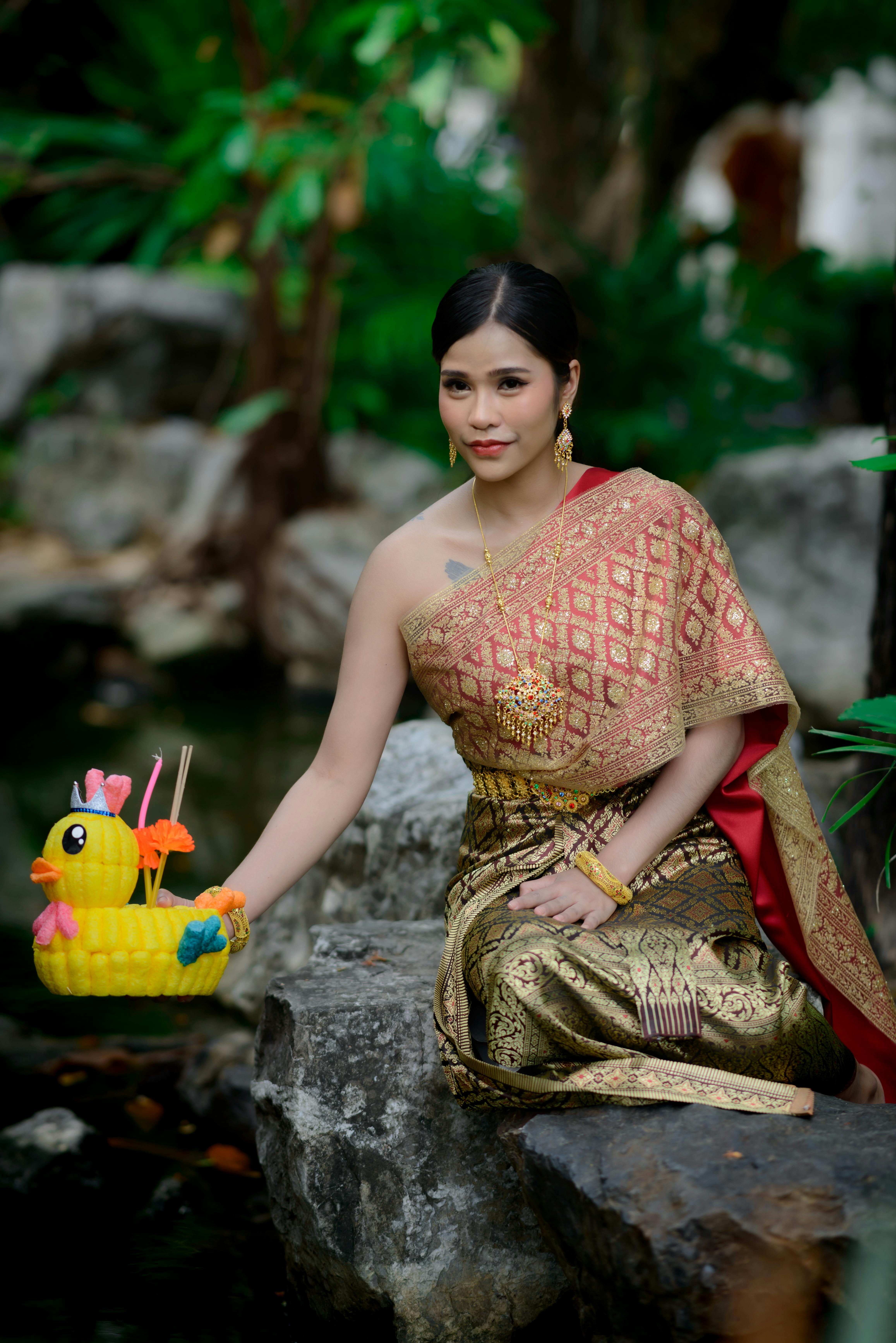 Asian woman in traditional dress holding a decorative basket with rubber duck by a pond.