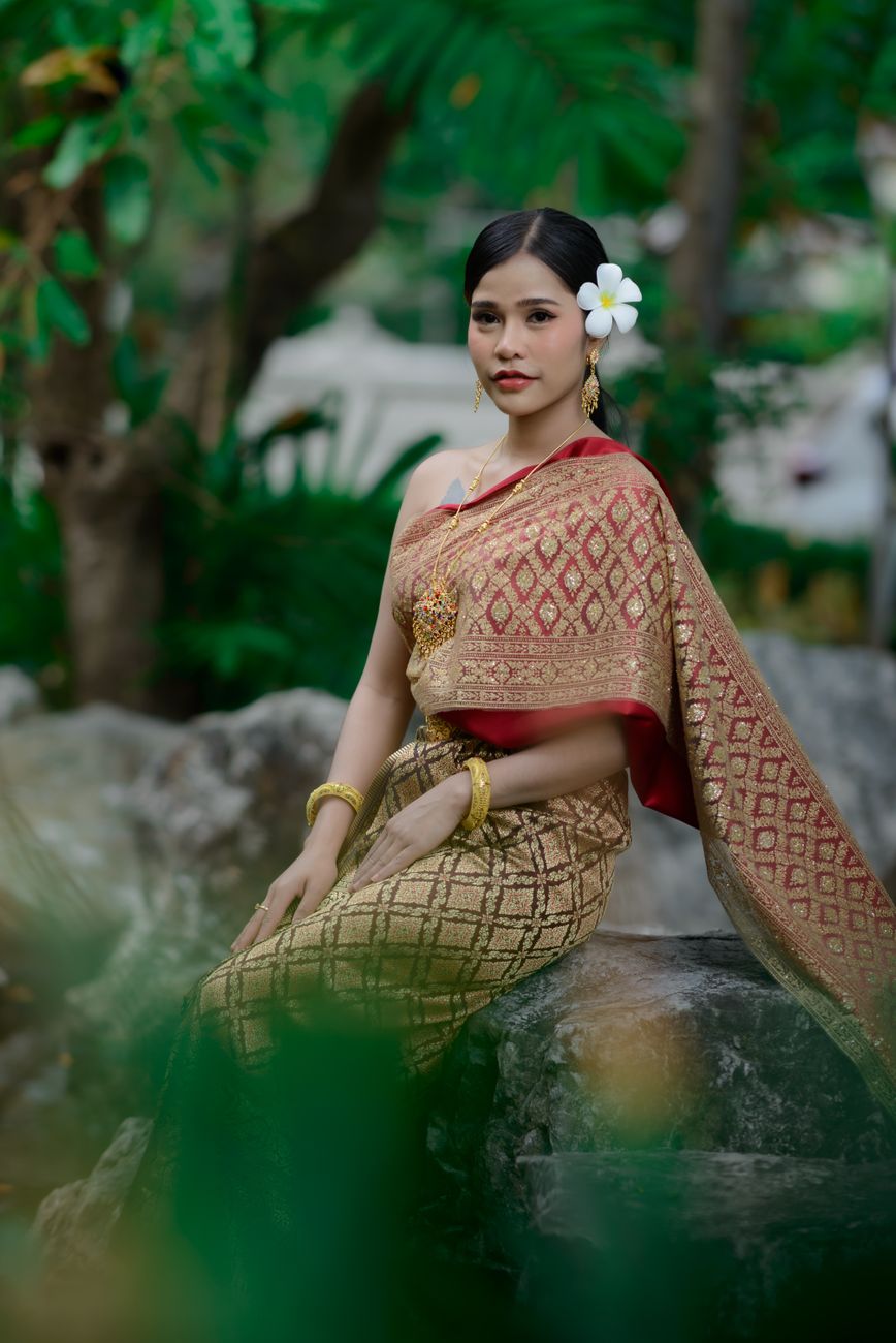 Asian woman in traditional Thai attire seated gracefully outdoors, exuding elegance.