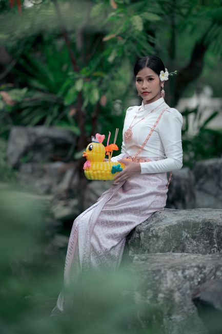 A woman in a traditional dress holds a toy in a lush garden setting, showcasing cultural attire.
