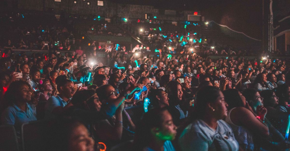 A crowded theater where audience members are holding up phones with voting screens, stage lit in neon colors