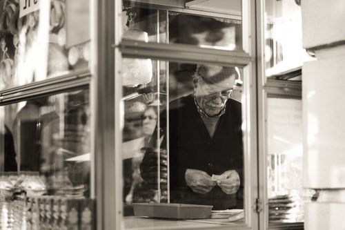 Free Black and white candid shot of a senior man at a kiosk in an urban setting, showcasing reflection and urban life. Stock Photo