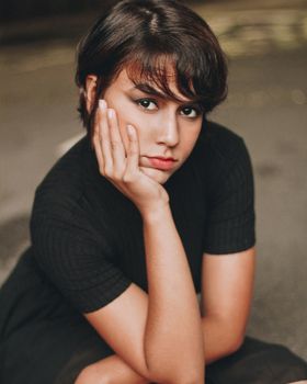 A thoughtful young woman with makeup rests her hand on her cheek in an outdoor setting.