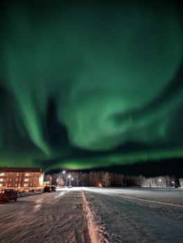 Stunning aurora borealis illuminates the night sky over a snow-covered road and buildings.