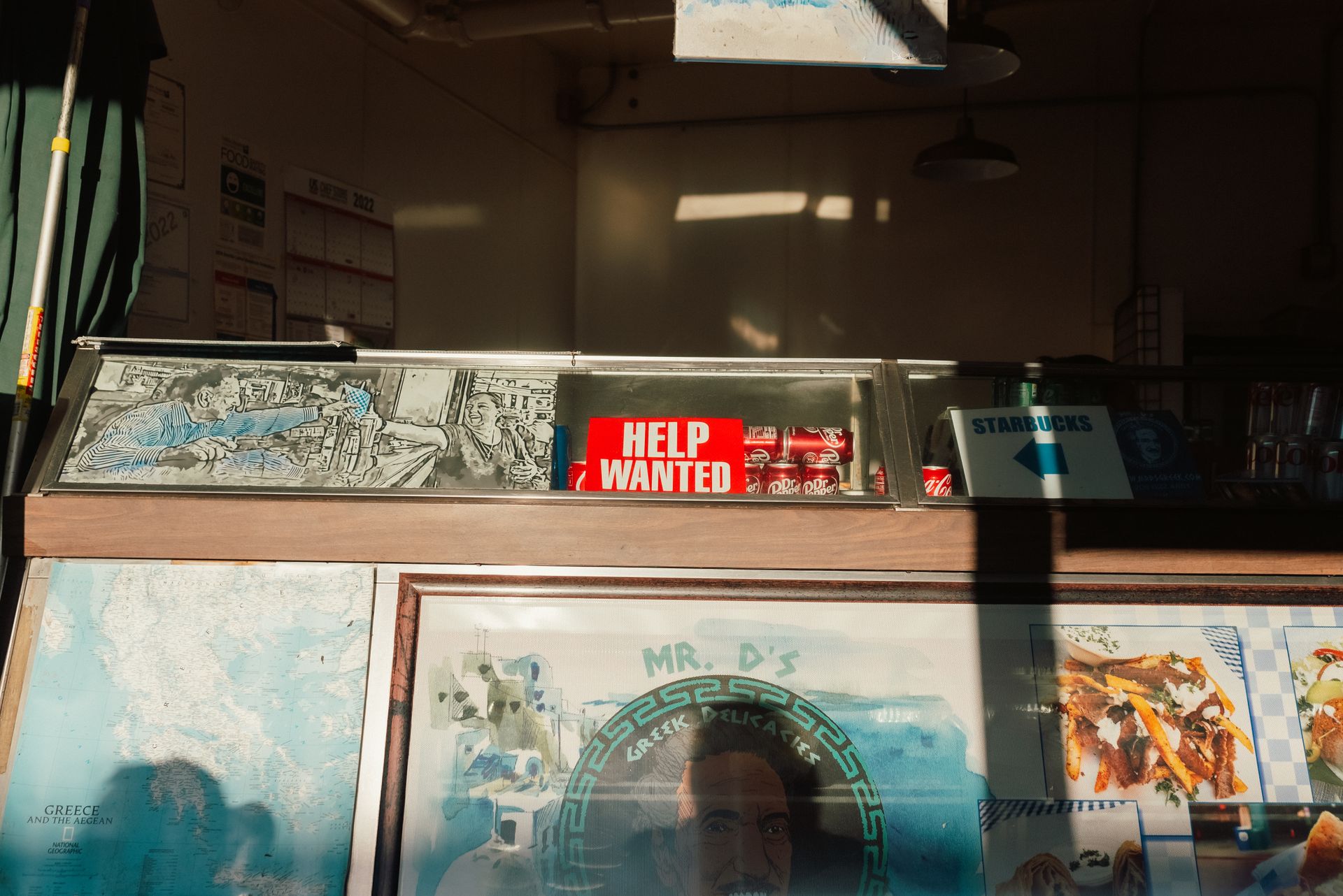 Image of a vintage deli interior with a prominent 'Help Wanted' sign and Greek decor.