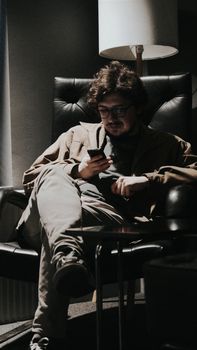 A man seated in a leather chair using his phone in a dimly lit room.