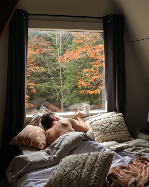 Man enjoying a relaxing morning in bed with a view of autumn foliage in Catskill, NY.