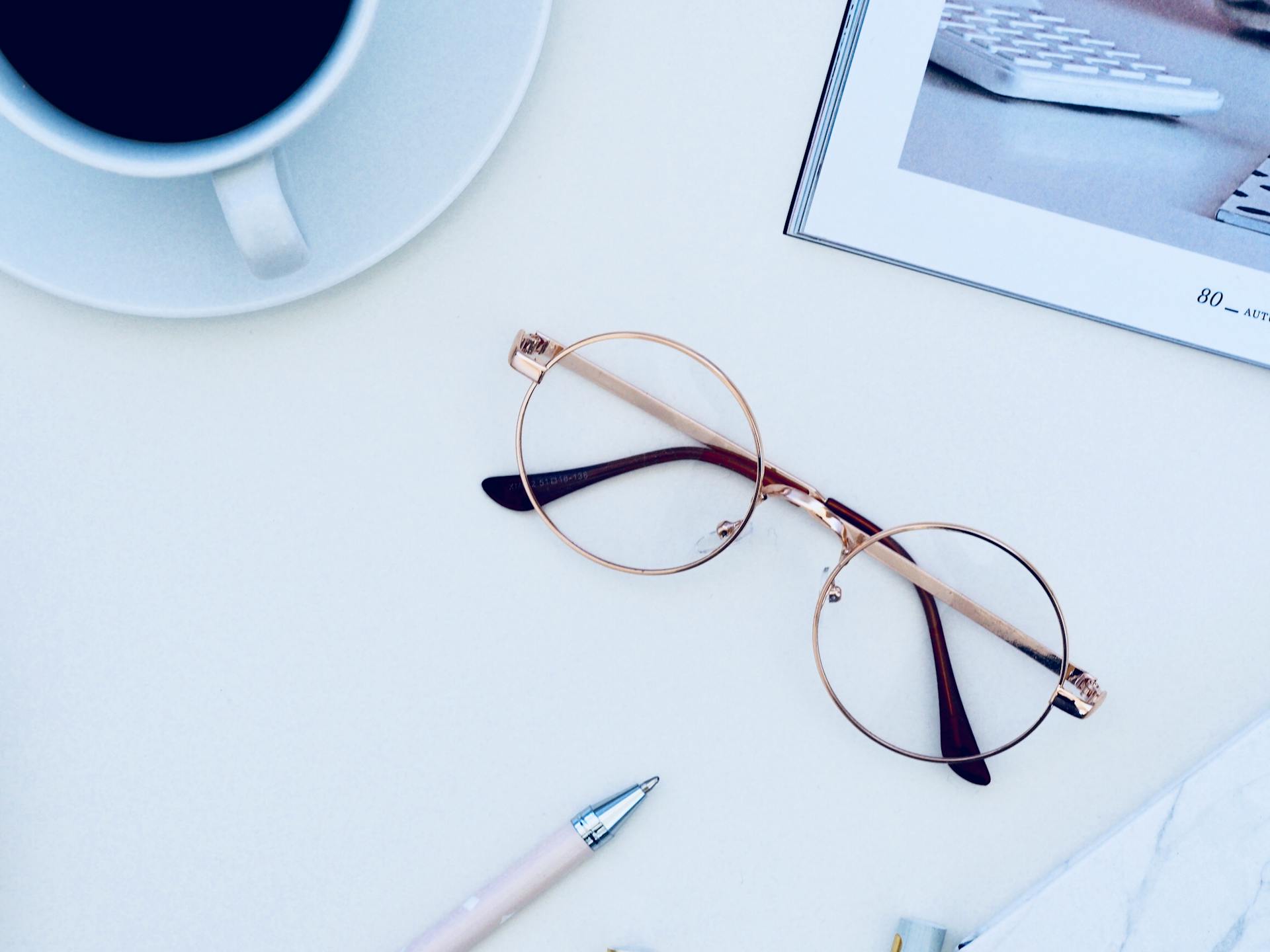 A clean, minimalist desk setup with a cup of coffee, eyeglasses, and a pen on a white surface.