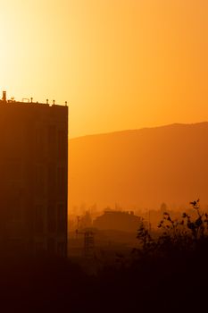 A backlit silhouette of buildings against a foggy sunset in Izmir, Turkey, with a warm, golden hue.