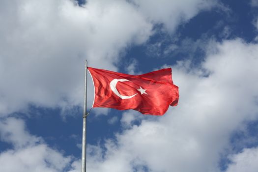 The Turkish flag waves proudly on a flagpole against a bright blue sky with clouds.