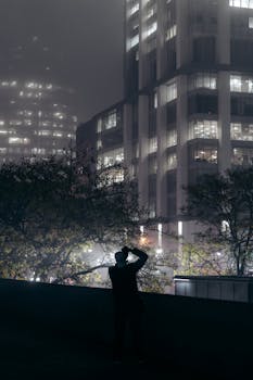 Silhouette of a photographer capturing skyscrapers amidst night fog in a cityscape.