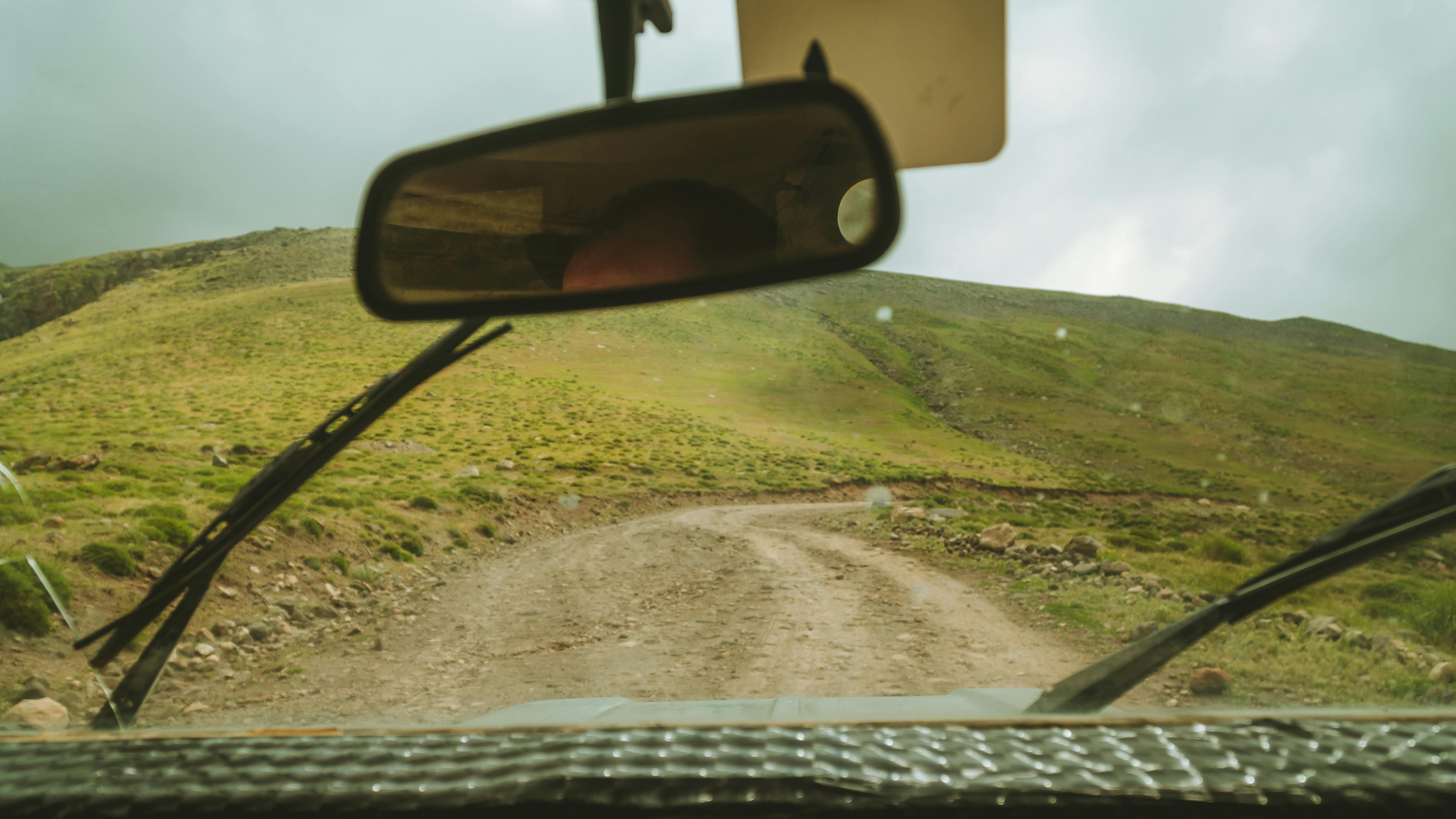 View from a vehicle's rearview mirror while off-roading on a rugged dirt path in a hilly landscape.