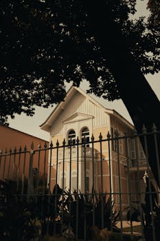 A beautiful Victorian-style house with ornate architectural details, partially obscured by a tree and iron fence.