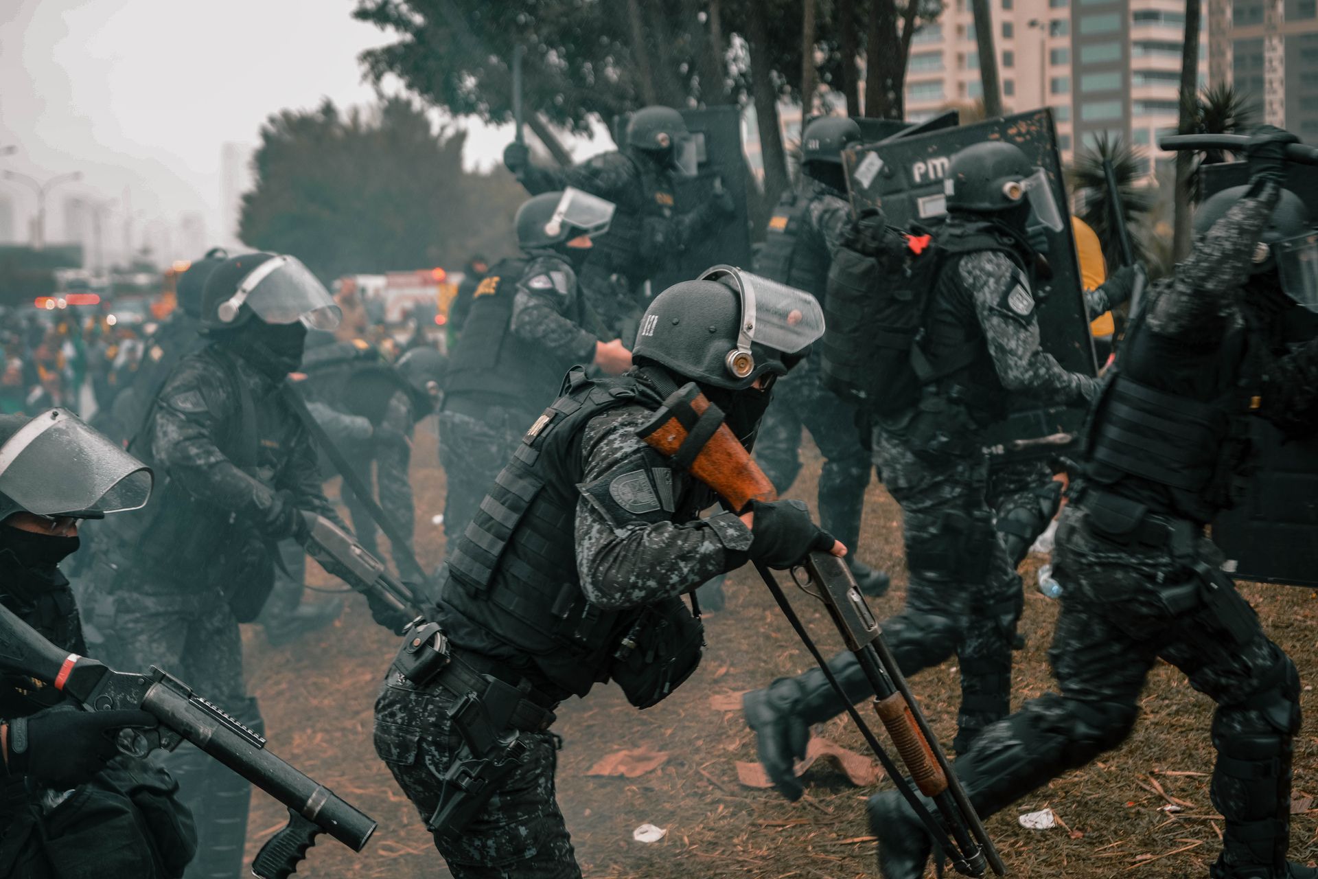Riot police equipped with shields and rifles during a protest in an urban setting.