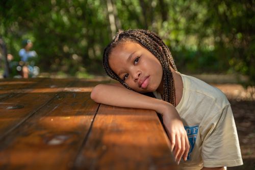 Free A young girl with braided hair leans on a wooden picnic table in a sunlit outdoor setting. Stock Photo