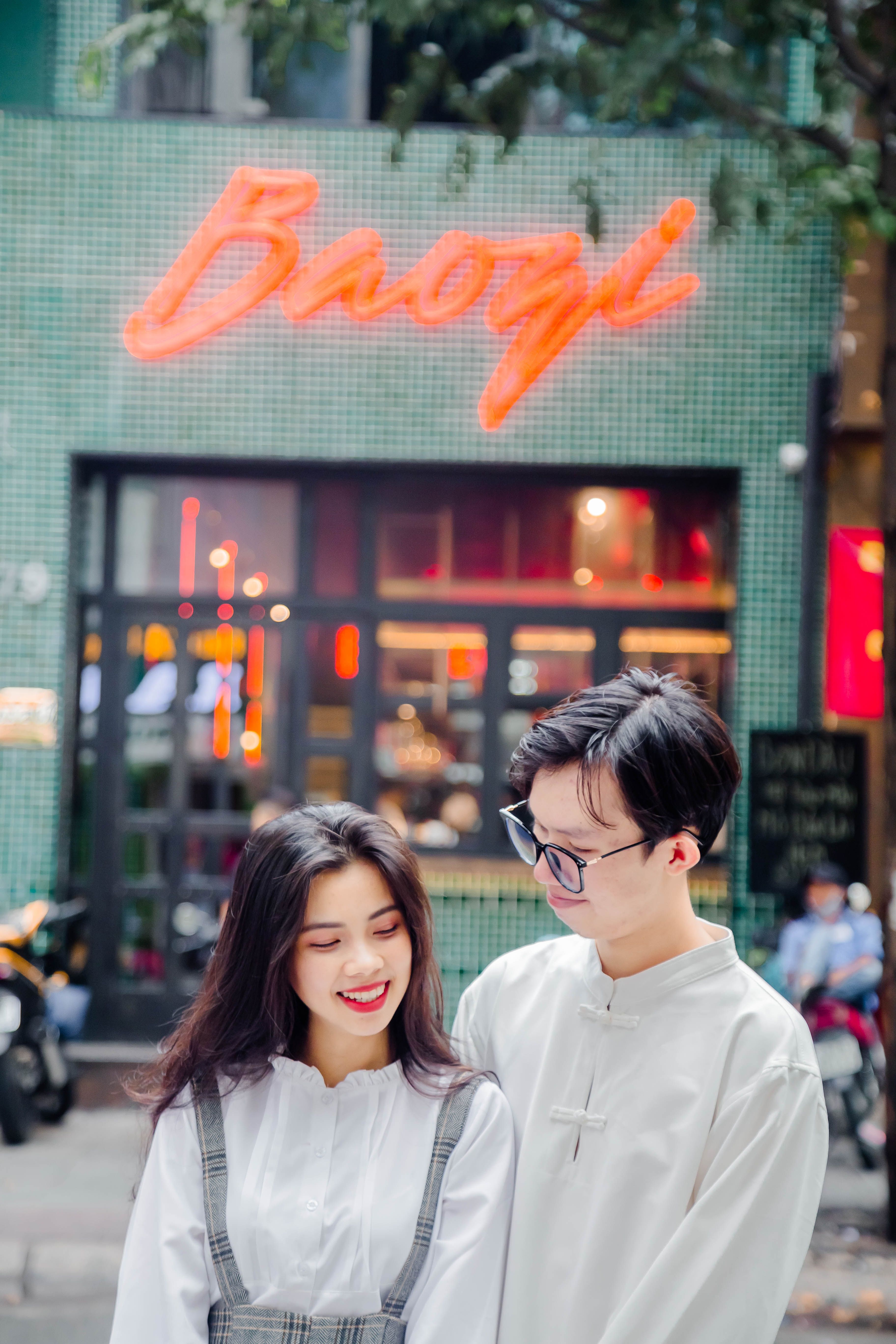 Young Asian couple smiling together in front of a neon-lit Baozi restaurant facade.