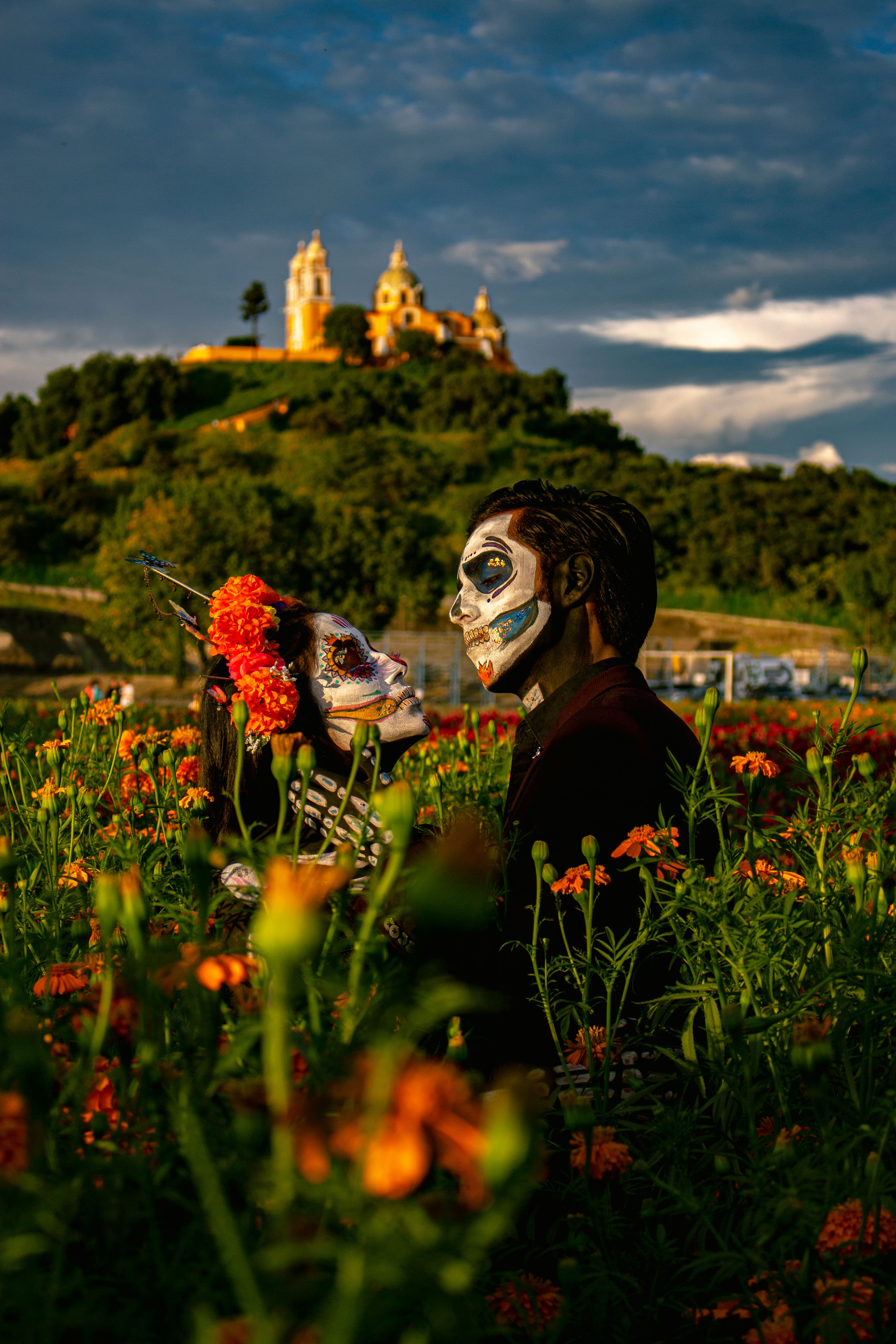 A couple with painted faces pose amidst vibrant flowers in Cholula, Mexico, celebrating Dia de Muertos.