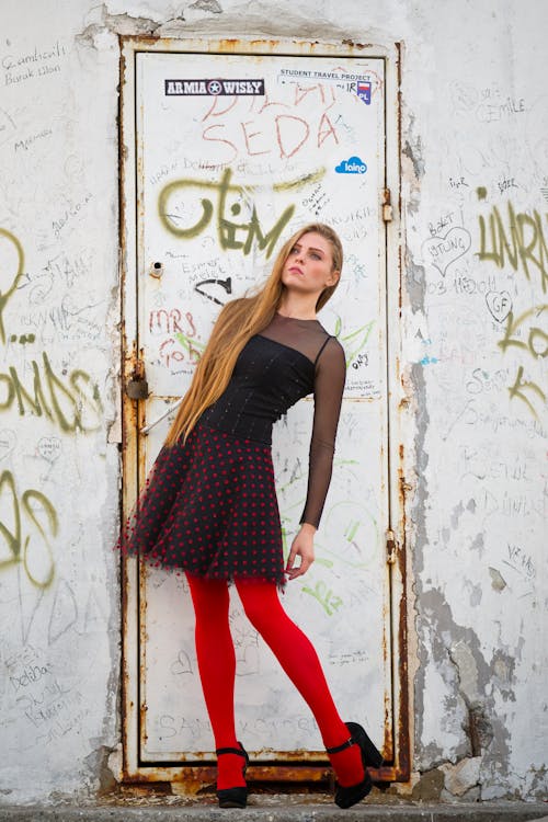 Free Woman Wearing Black and Red Long-sleeved Mini Dress and Red Leggings in Front of a Closed White Metal Door Stock Photo